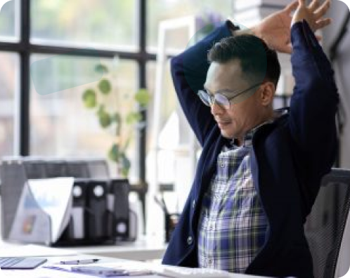 A man with glasses wearing a plaid shirt and blue jacket sitting at his office desk and stretching his arms behind his head during a work break.