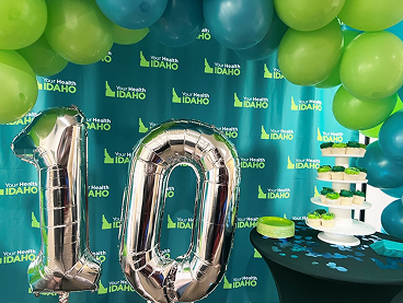 Large silver foil balloons forming the number 10 standing in front of a teal "Your Health Idaho" step-and-repeat backdrop decorated with green and blue balloon arches and a tiered cupcake stand.