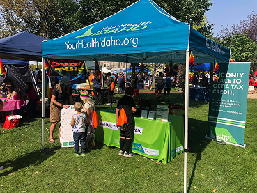 A teal "Your Health Idaho" branded popup canopy tent set up on a grassy lawn at a community event, featuring a green table banner, promotional signs, and attendees interacting with event staff.