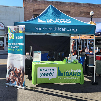 A teal "Your Health Idaho" branded popup canopy tent and promotional banner setup at an outdoor street event, featuring a green tablecloth display with the slogan "HEALTH, yeah!" and informational materials.